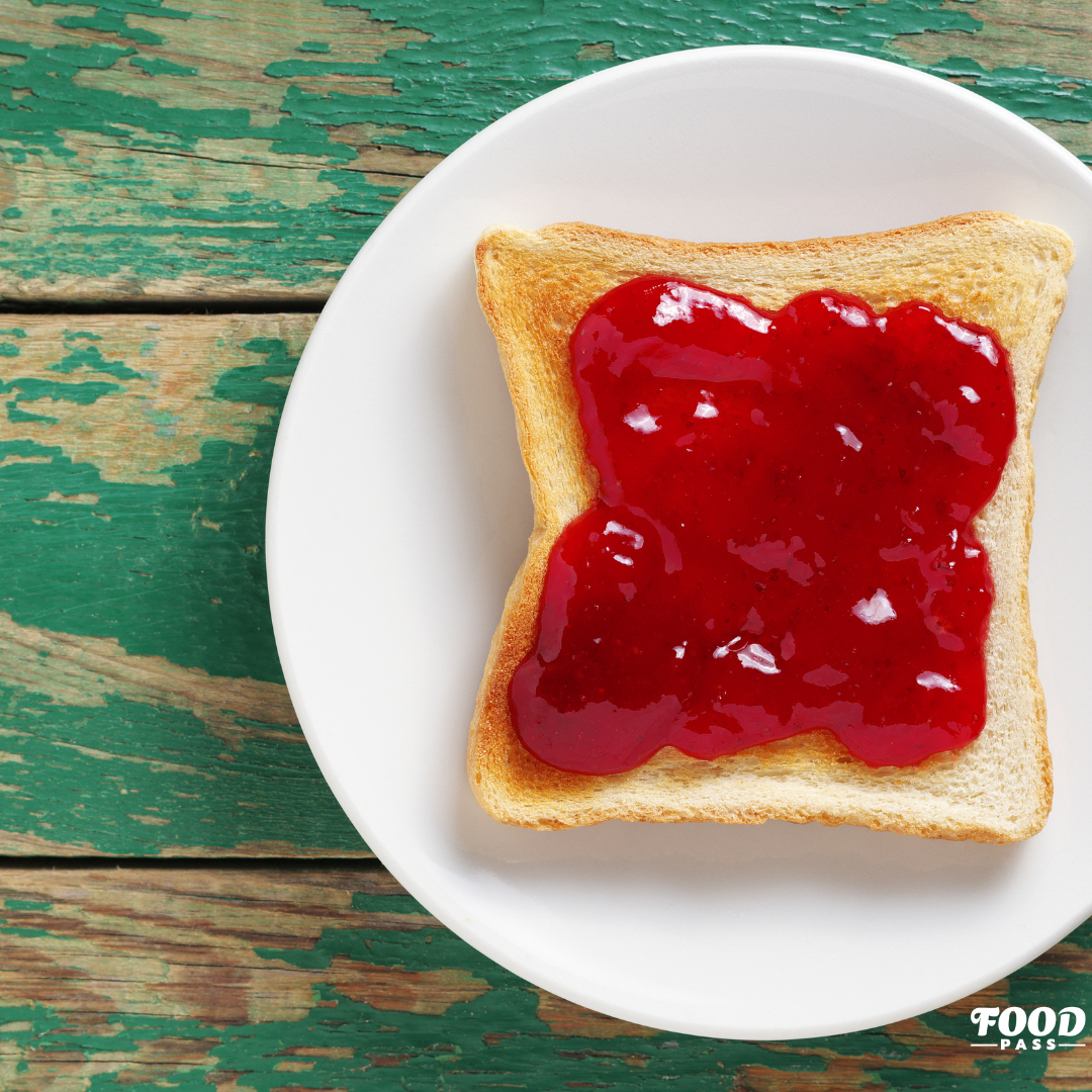 Tranche de pain grillé garnie d’une confiture rouge éclatante sur une assiette blanche, posée sur une table en bois rustique. Une mise en avant des confitures artisanales Simon Turcotte Confiturier, produit vedette de la boîte FoodPass de février.